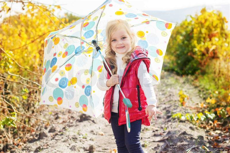 Smiling little girl with colorful umbrella is wearing red vest and boots walking on the vineyard, stock photo