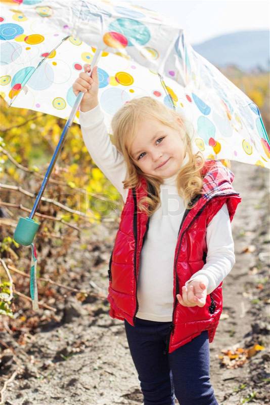 Smiling little girl with umbrella is wearing red vest and boots walking on the vineyard, stock photo