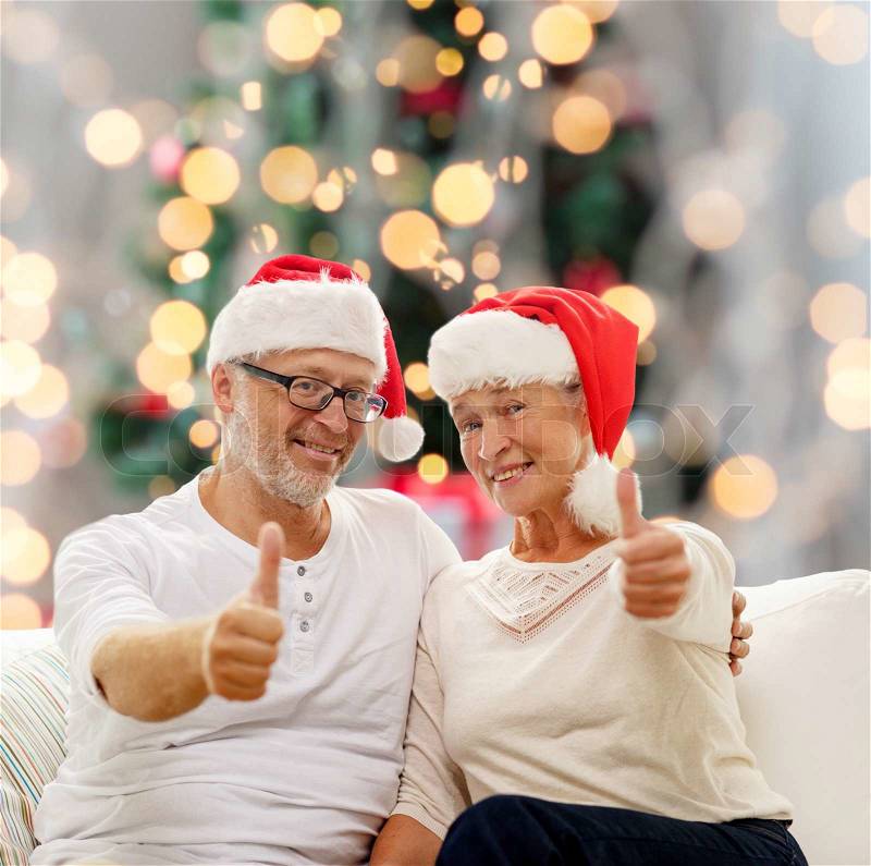 Family, holidays, age and people concept - happy senior couple in santa helper hats sitting on sofa over christmas tree lights background, stock photo