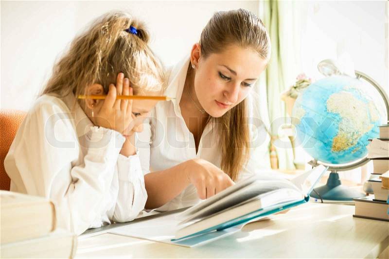Young mother and daughter reading textbook while doing homework, stock photo