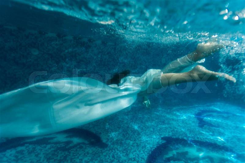 Underwater shot of beautiful woman wearing white fabric diving in pool, stock photo