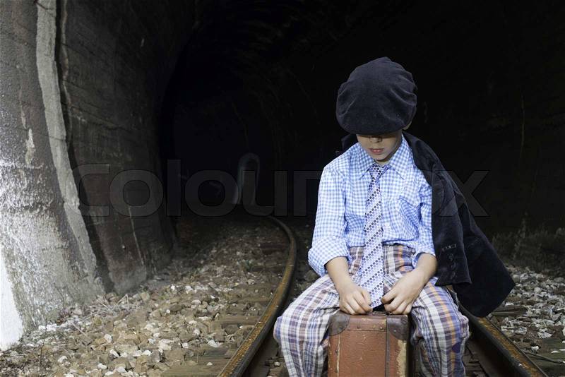 Child in vintage clothes sits on railway road in front of a tunnel, stock photo
