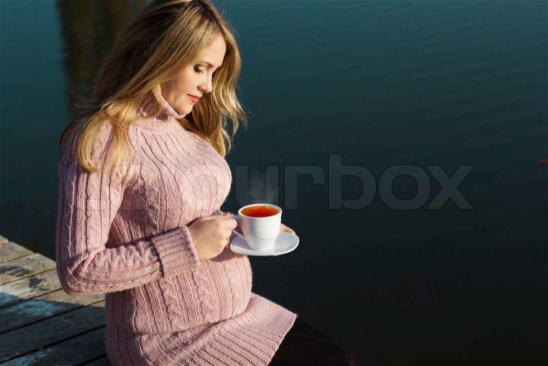 Pregnant beautiful adult woman is drinking hot tea near autumn lake with pink ribbon on her belly. Love and care concept, stock photo