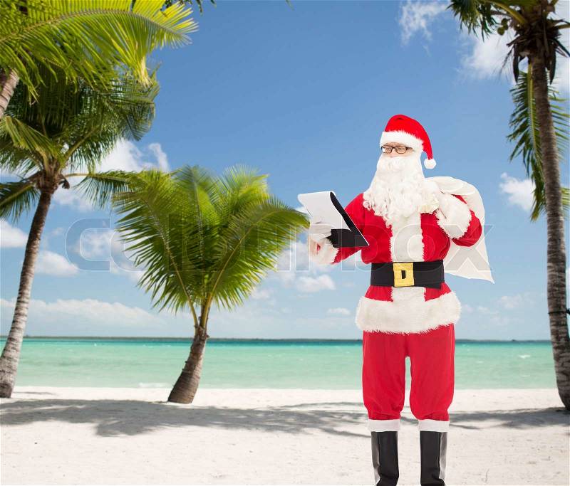 Christmas, holidays and people concept - man in costume of santa claus with notepad and bag over tropical beach background, stock photo