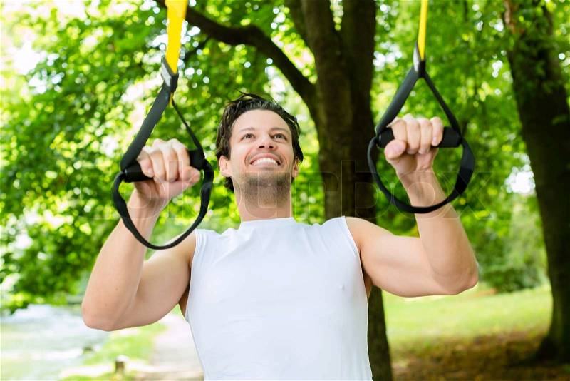 Young man exercising with suspension trainer sling in City Park under summer trees for sport fitness, stock photo