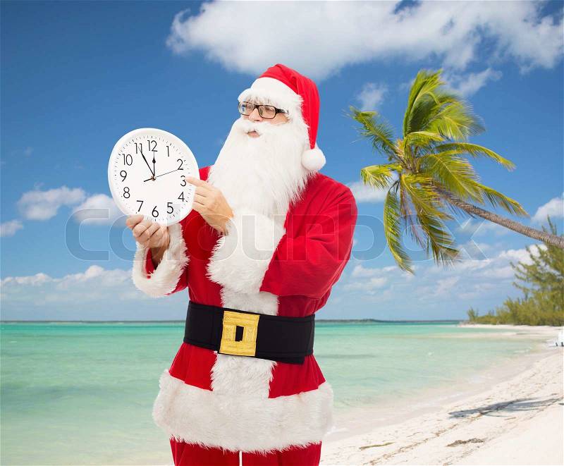 Christmas, holidays and people concept - man in costume of santa claus with clock showing twelve pointing finger over tropical beach background, stock photo