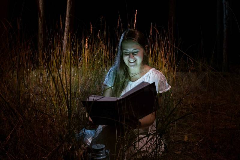 Portrait of happy woman holding glowing book at night forest, stock photo