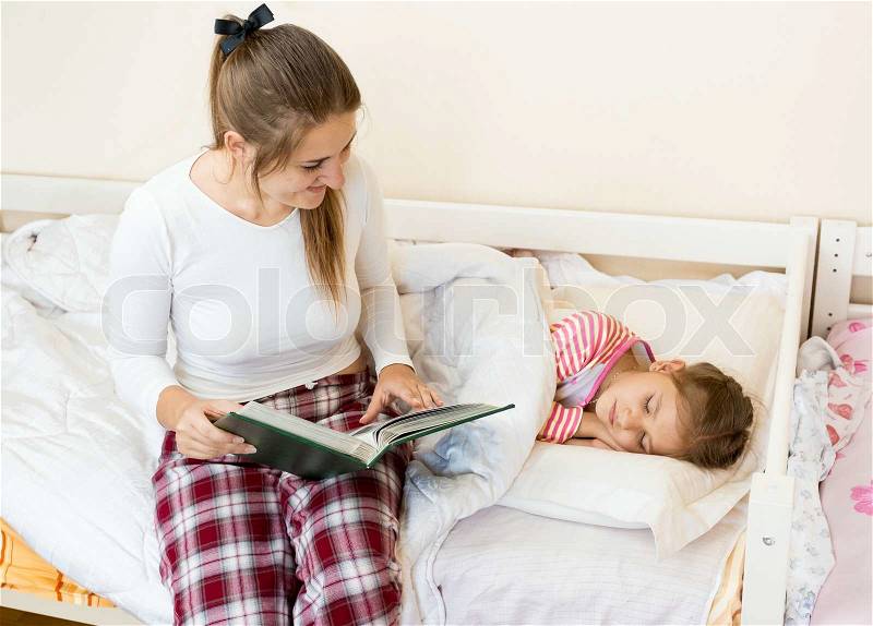 Young mother sitting on bed next to daughter and reading a story, stock photo