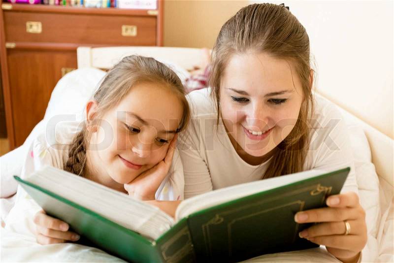 Portrait of smiling mother and daughter reading big book at bed, stock photo