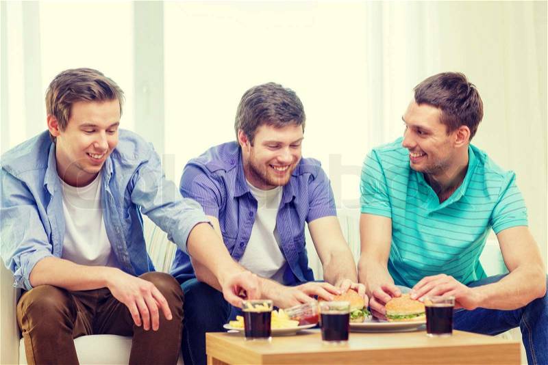 Friendship, food and leisure concept - smiling friends with soda and hamburgers at home, stock photo