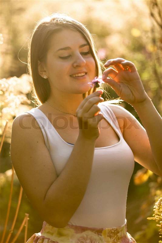 Toned portrait of smiling young woman tearing petal off flower at field, stock photo