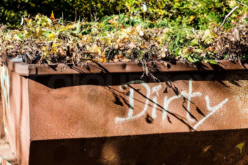 Old rusty metal waste container with green garden waste and word dirty painted on it, stock photo