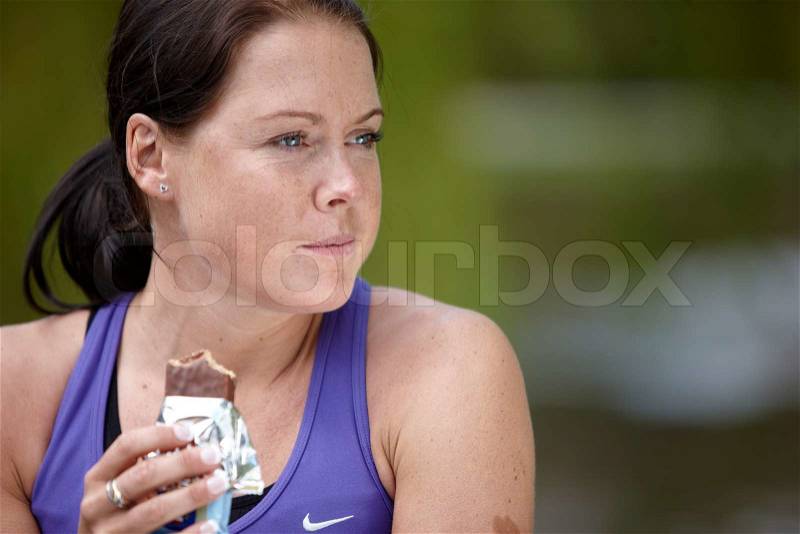 A young brunette female in violet sportswear eating chocolate energy bar, stock photo