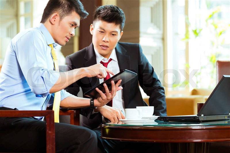 Two Asian Chinese businessman or office people having a business meeting in a hotel lobby discussing documents on a tablet computer while drinking coffee , stock photo