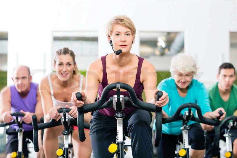 Senior and young people spinning on fitness bike in gym doing endurance and cardio training, the instructor is leading them on, stock photo