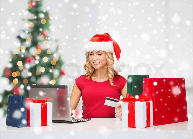 Technology, holidays, online shopping and people concept - woman in santa helper hat with gift boxes and laptop computer over living room background, stock photo
