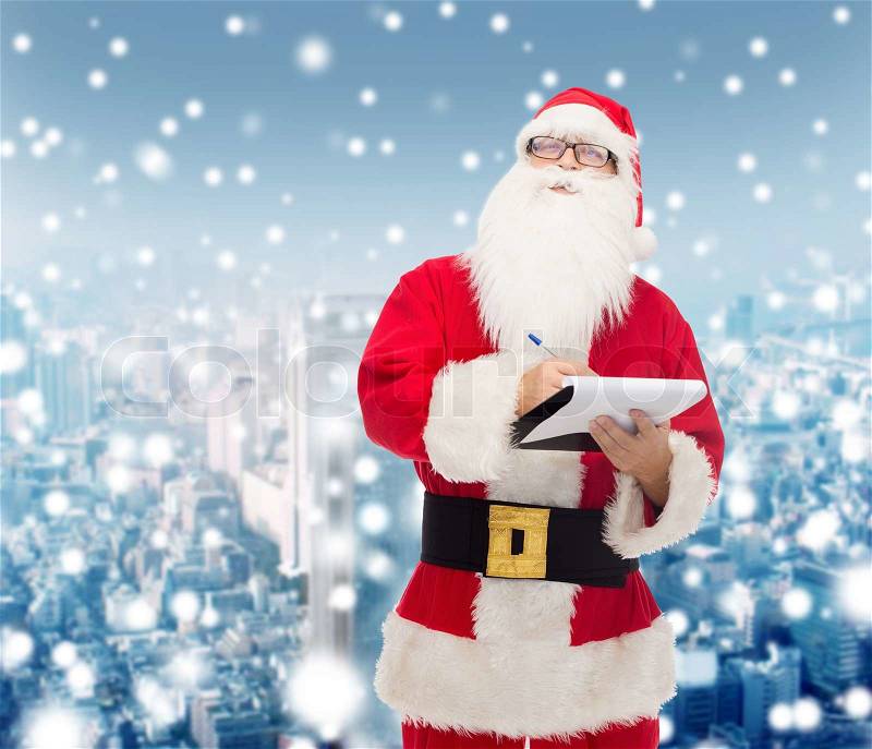 Christmas, holidays and people concept - man in costume of santa claus with notepad and pen over snowy city background, stock photo
