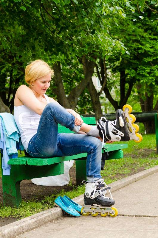 Attractive blond woman sitting on a park bench putting on roller blades as she prepares for a fun day skating, stock photo