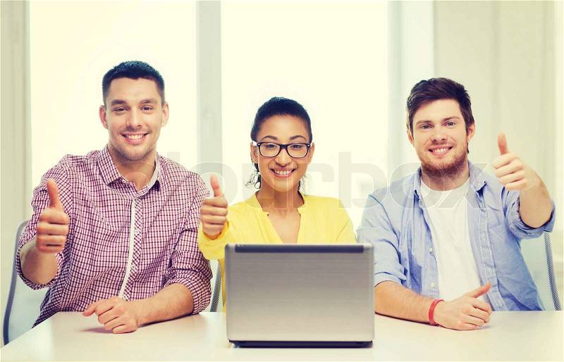 Education, technology, business, startup and office concept - three smiling colleagues with laptop in office showing thumbs up, stock photo