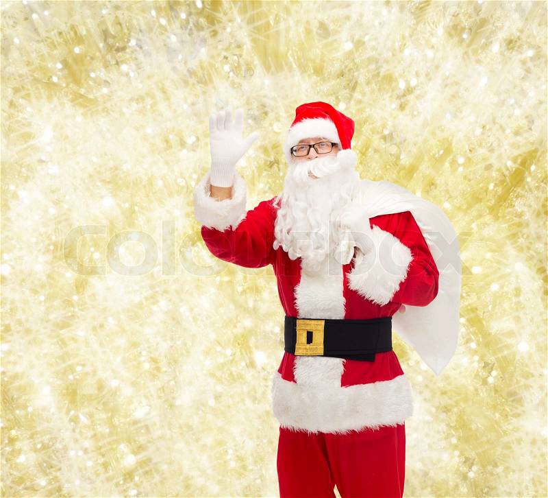 Christmas, holidays, gesture and people concept - man in costume of santa claus with bag waving hand over yellow lights background, stock photo