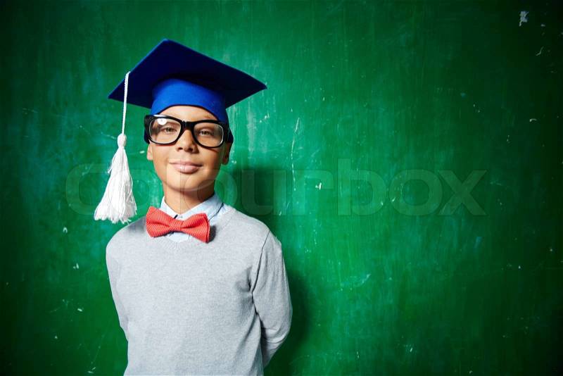 Elementary learner in eyeglasses and graduation hat posing in isolation, stock photo