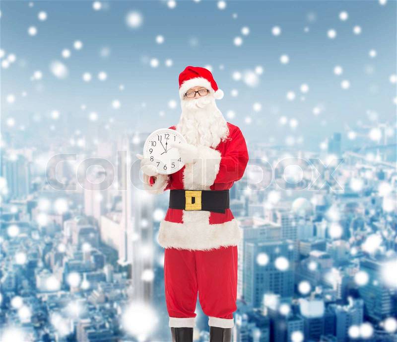 Christmas, holidays and people concept - man in costume of santa claus with clock showing twelve pointing finger over snowy city background, stock photo