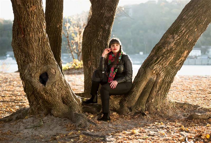 Young woman sitting under tree at autumn park next to river, stock photo