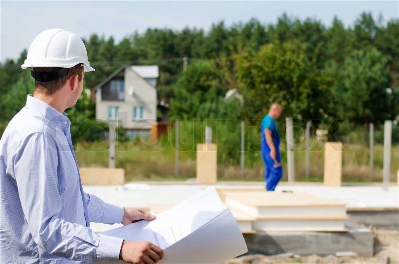 Architect or engineer checking plans on site holding the open blueprint in his hands as builders work in the background, stock photo