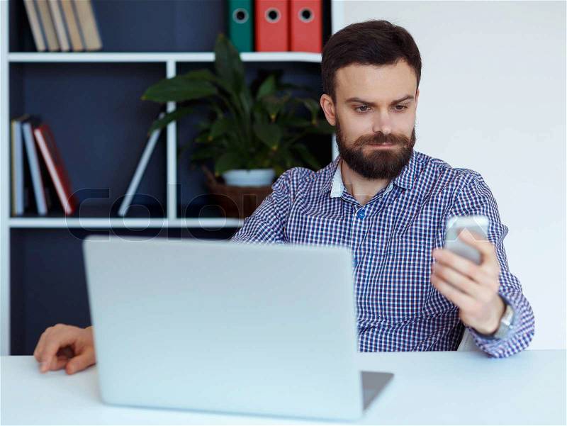 Young bearded man working on a laptop in the office and talking on cell phone, stock photo