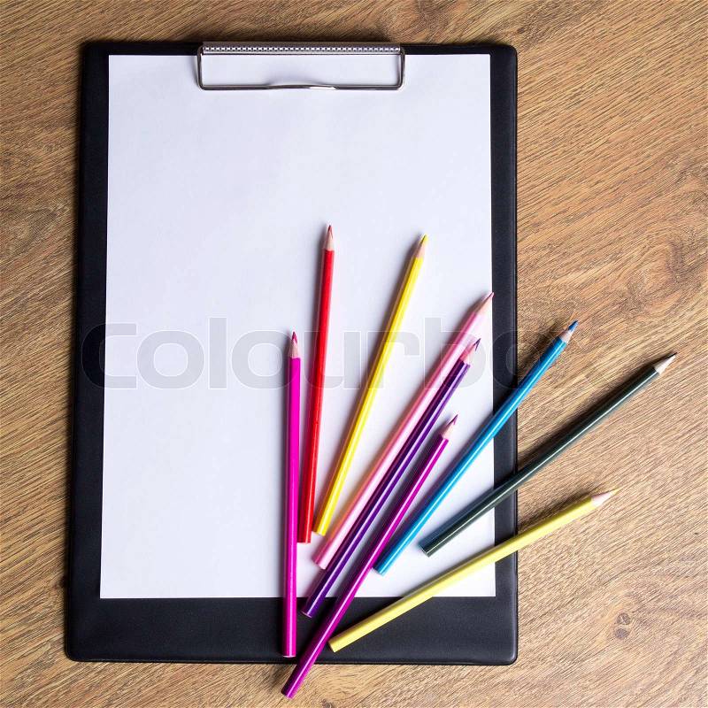 Colored drawing pencils and clipboard with blank paper on wooden table background, stock photo