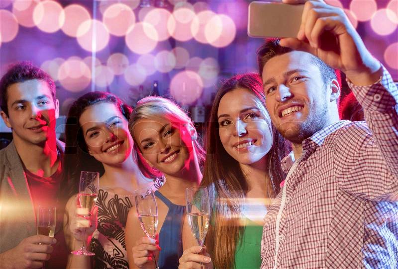 Party, holidays, technology, nightlife and people concept - smiling friends with glasses of champagne and smartphone taking selfie in club, stock photo