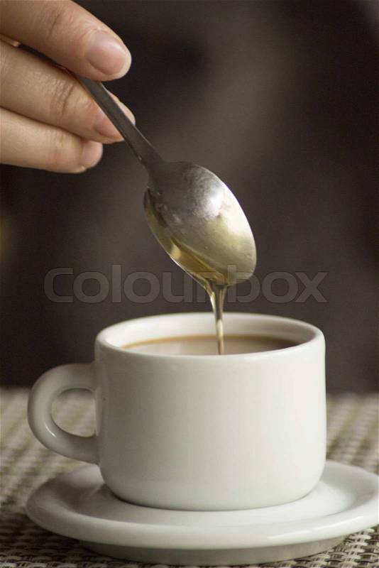 Hand of young woman ading corn syrup with spoon in cup of coffee expresso with milk on saucer in table, stock photo
