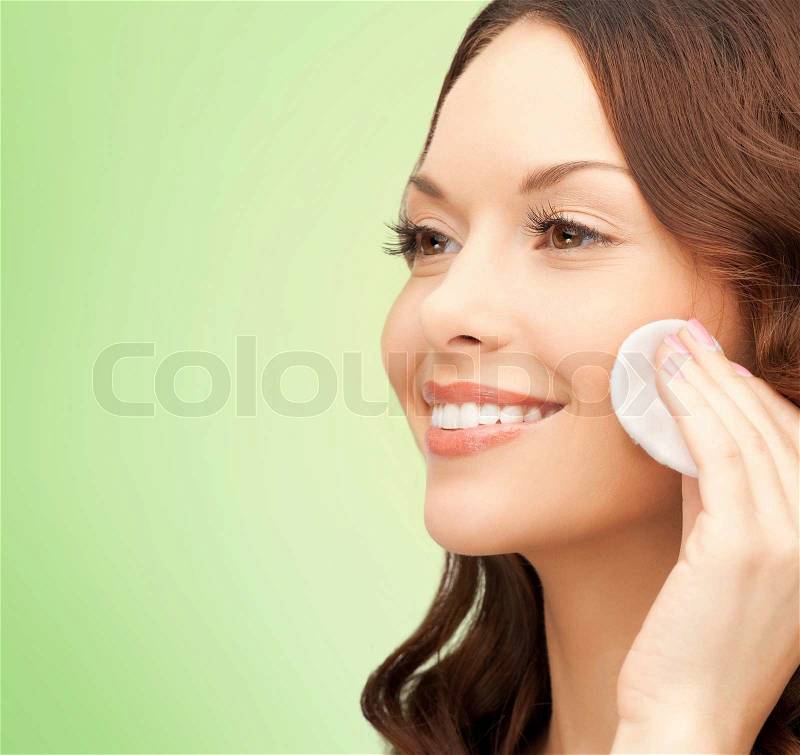 Beauty, people and health concept - beautiful smiling woman cleaning face skin with cotton pad over green background, stock photo