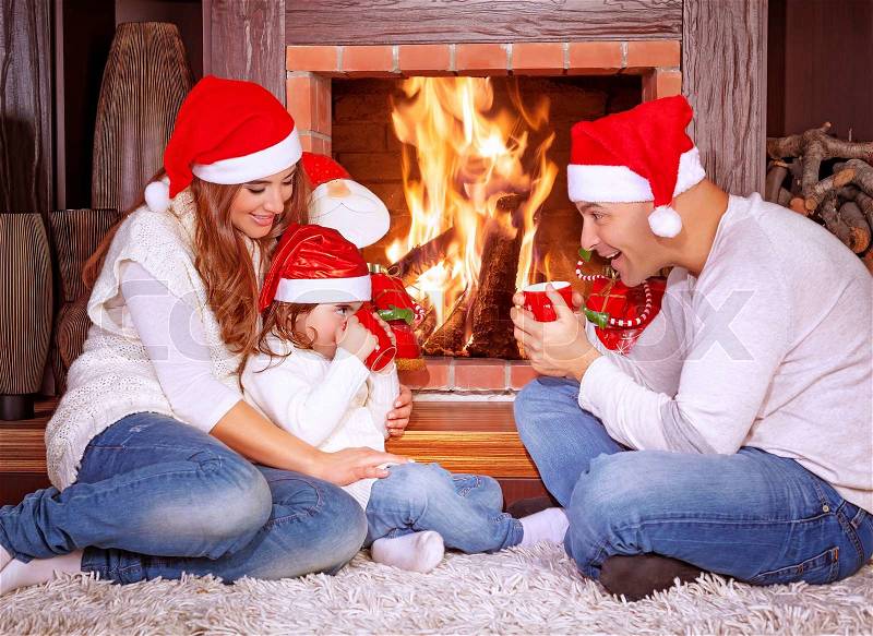 Happy family by fireplace, parents with baby girl wearing Santa Claus hats, sitting relaxed in ski resort chalet and drinking hot tea, joyful winter vacation, people traveling on Christmas holidays, stock photo