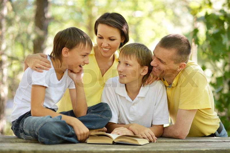 Family of four reading outdoors at table with book, stock photo