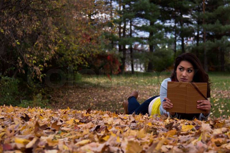 Young woman reading a book lying down on autumn leaves in the fall, stock photo