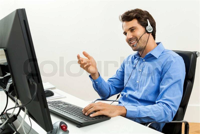 Attractive unshaven young man wearing a headset offering online chat and support on a client services of help desk as he types in information on his computer, stock photo