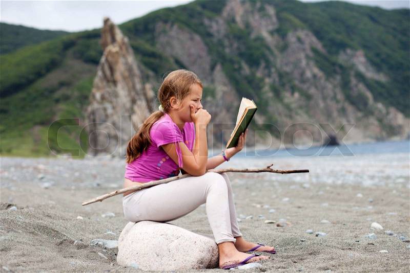 Girl reading book sitting on a stone on a sandy sea shore with rocka on the background, stock photo