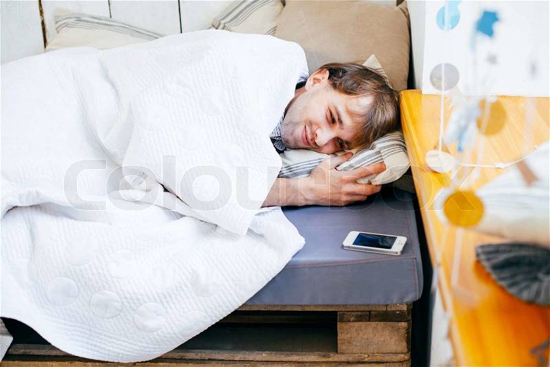 Sleepy young man in bed extending hand to alarm clock, stock photo