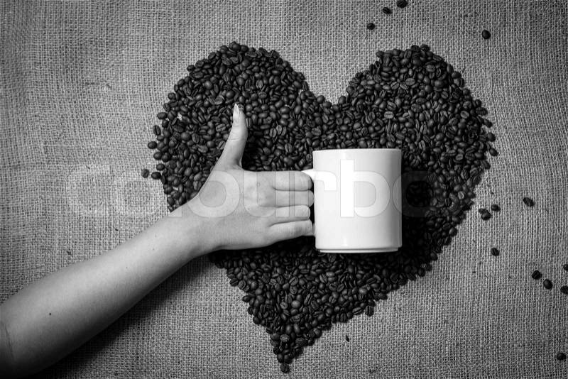 Black and white photo of hand with thumb up holding mug against heart made of coffee beans, stock photo