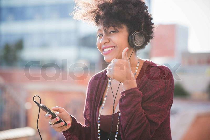 Beautiful black curly hair african woman listening music with headphones in town, stock photo