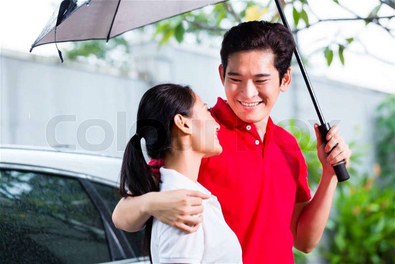 Asian man with umbrella walking woman in rain from car door to house, stock photo
