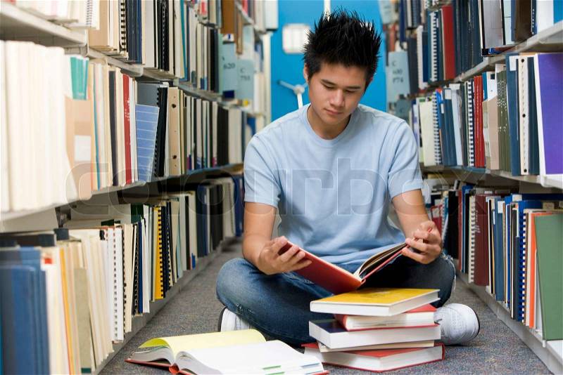 Male university student sitting on library floor surrounded by books, stock photo