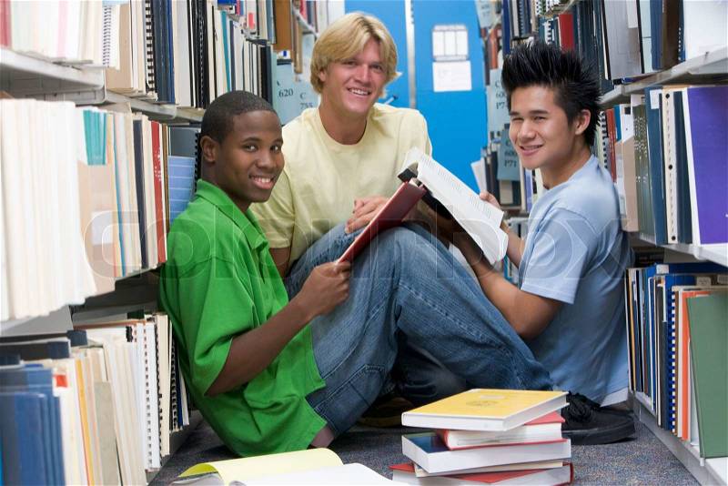 Group of three male students sitting on floor of library surrounded by books, stock photo