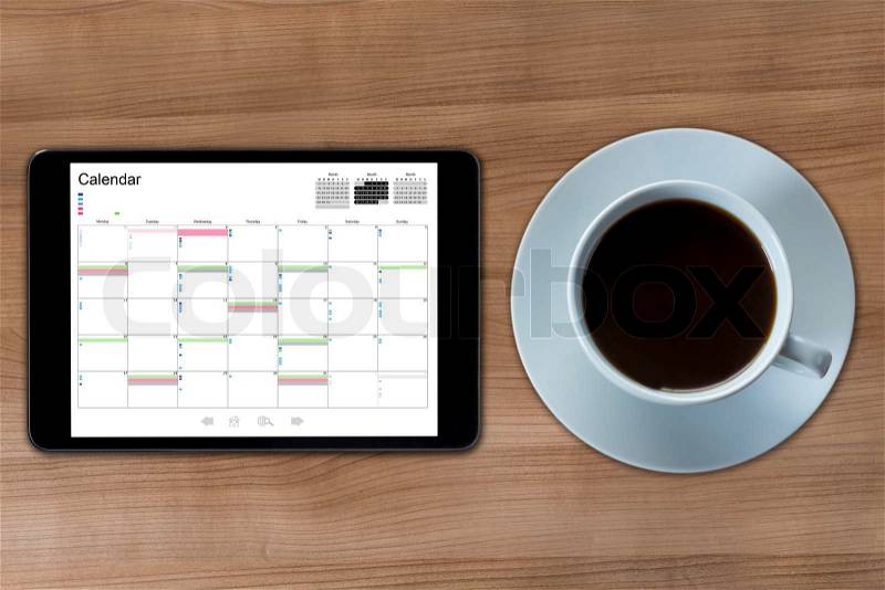 A tablet computer with a calendar displayed and a cup of coffee on a wooden desktop, stock photo