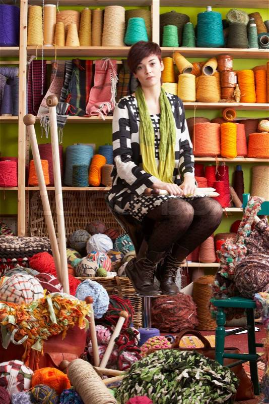 Young Woman Sitting On Stool Holding Knitting Needles In Front Of Yarn Display, stock photo