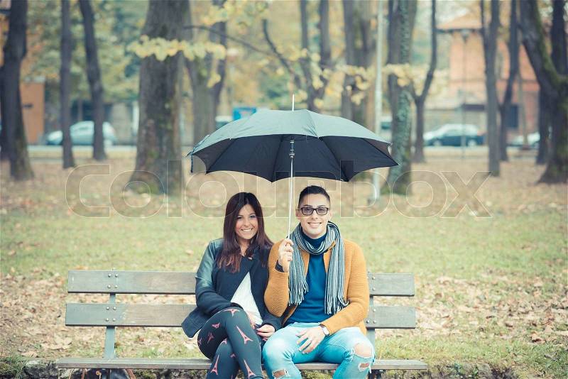 Young couple in the park during autumn season outdoor - lovers valentine under umbrella sitting in a bench, stock photo