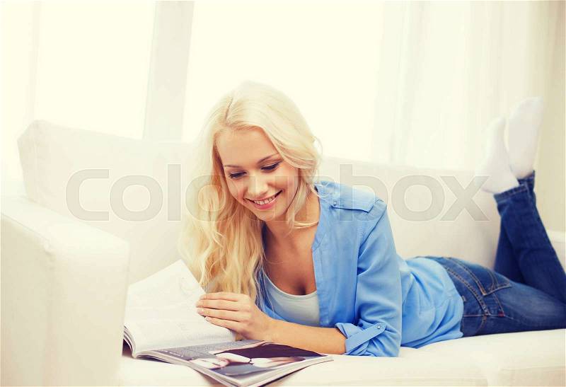 Home and leasure concept - smiling woman lying on couch and reading magazine at home, stock photo