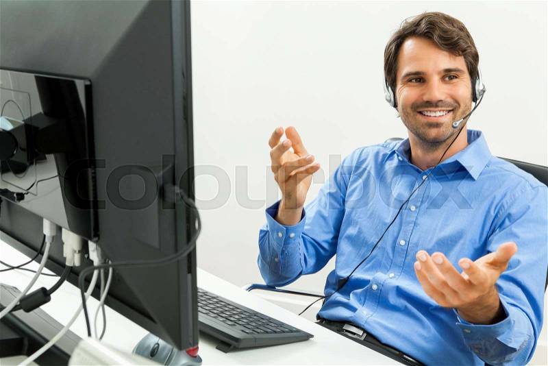 Attractive unshaven young man wearing a headset offering online chat and support on a client services of help desk as he types in information on his computer, stock photo
