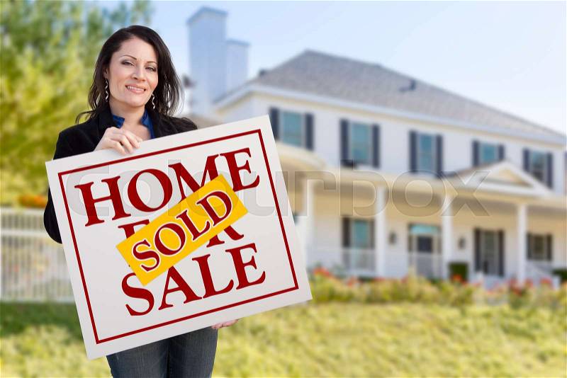 Smiling Hispanic Woman Holding Sold Home For Sale Sign In Front of Beautiful House, stock photo